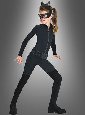 Ein Mädchen im Catwoman-Kinderkostüm – bestehend aus einem schwarzen Catsuit, original lizenzierten Katzenohren (Katzenohren-Stirnband), einer Augenmaske und schwarzen Stiefeln – nimmt vor einem grauen Hintergrund eine geduckte, kampfbereite Haltung ein und verkörpert damit perfekt den ikonischen Catwoman-Look.