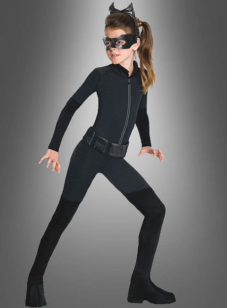 Ein Mädchen im Catwoman-Kinderkostüm – bestehend aus einem schwarzen Catsuit, original lizenzierten Katzenohren (Katzenohren-Stirnband), einer Augenmaske und schwarzen Stiefeln – nimmt vor einem grauen Hintergrund eine geduckte, kampfbereite Haltung ein und verkörpert damit perfekt den ikonischen Catwoman-Look.