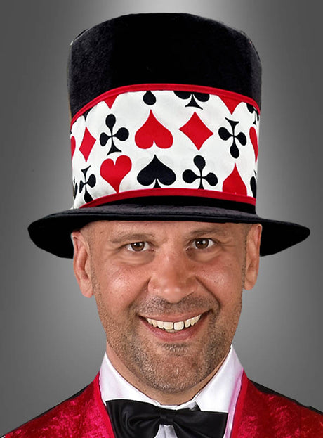 Ein Mann trägt den „Casino Hut Stoff Zylinder Unisex“, einen hohen Hut, der mit Spielkartenfarben verziert ist, dazu eine rote Samtjacke, ein weißes Hemd und eine schwarze Fliege, und lächelt vor einem schlichten grauen Hintergrund.