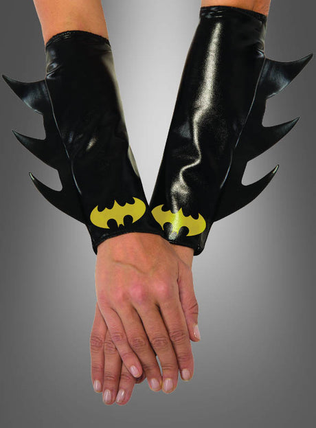 Vor einem grauen Hintergrund mit Farbverlauf sind zwei Hände übereinandergelegt, an denen jeweils Batgirl-Armstulpen zu sehen sind – glänzende schwarze Manschetten mit Stacheln in Form von Fledermausflügeln und leuchtend gelben Batman-Logos an den Handgelenken.