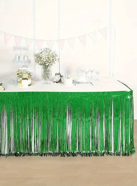 Ein für eine Party geschmückter Tisch wird durch den glänzend grünen „Lametta Tischrock“ (75 x 275 cm) als Tischumrandung geprägt, der mit Blumen, Glaswaren und einem Etagerenständer verziert ist. Eine weiße Wand und pastellfarbene Wimpelketten im Hintergrund runden die bezaubernde Partydekoration ab.