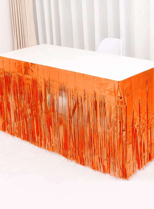 Ein weißer Tisch ist mit dem „Lametta Tischrock 75 x 275 cm“ in leuchtendem Orange geschmückt, was einem hellen Raum mit weißen Vorhängen und einem weißen Stuhl einen festlichen Partydekorations-Touch verleiht.