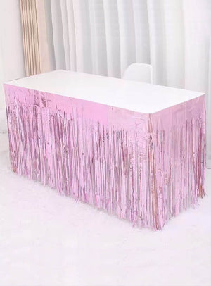 Ein rechteckiger Tisch mit weißer Tischplatte ist an den Seiten mit dem „Lametta Tischrock 75 x 275 cm“ in glänzendem Rosa verziert und steht vor weißen Vorhängen auf einem weißen Teppich, wodurch eine festliche Partydekoration entsteht.