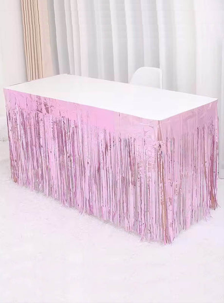 Ein rechteckiger Tisch mit weißer Tischplatte ist an den Seiten mit dem „Lametta Tischrock 75 x 275 cm“ in glänzendem Rosa verziert und steht vor weißen Vorhängen auf einem weißen Teppich, wodurch eine festliche Partydekoration entsteht.