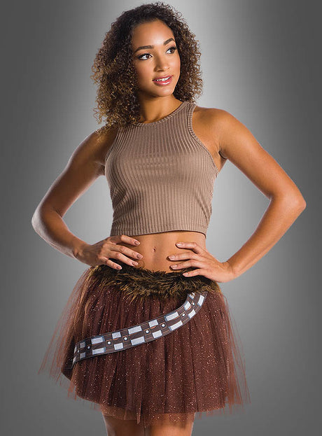 Eine Frau präsentiert ein ärmelloses, beiges Oberteil in Kombination mit dem Chewbacca-Wookiee-Tüllrock – einem braunen Tüllrock mit Pelzbesatz und gemusterter Schärpe – und posiert mit den Händen in den Hüften vor einem grauen Hintergrund mit Farbverlauf. Dieses Ensemble ist eine schicke, dezente Interpretation des legendären Star-Wars-Wookiee-Damenkostüms.