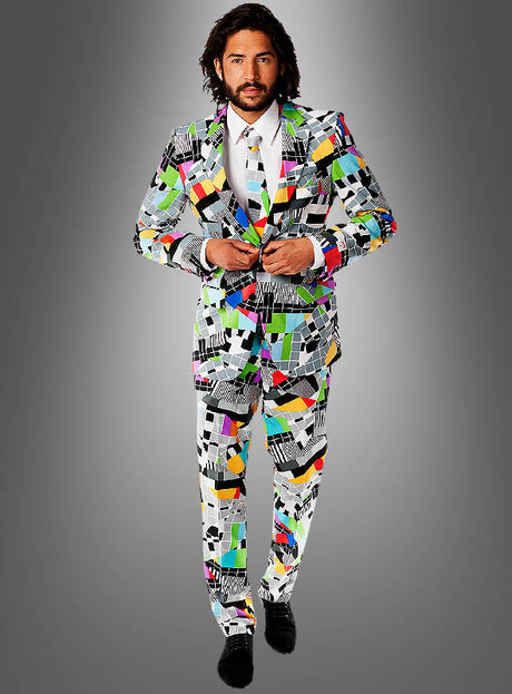 Ein Mann, der den „Deluxe Testbild Anzug“ von OppoSuits trägt – einen farbenfrohen Anzug mit geometrischem Muster, dazu ein weißes Hemd und eine Krawatte –, steht vor einem grauen Hintergrund mit Farbverlauf und ist damit die perfekte Wahl für eine 70er-Jahre-Mottoparty oder als auffälliges Karnevalskostüm.
