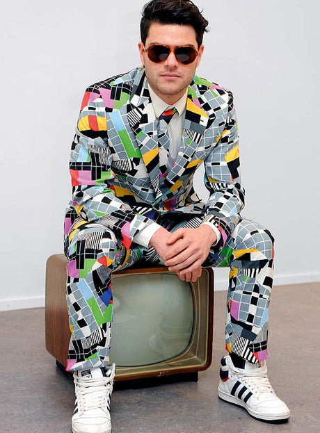 Ein Mann in einem farbenfrohen „Deluxe Testbild“-Anzug von OppoSuits mit geometrischem Muster und Sonnenbrille sitzt auf einem alten Fernseher und trägt weiße Sneaker vor einem schlichten Hintergrund – ideal für eine 70er-Jahre-Mottoparty oder als auffälliges Karnevalskostüm.