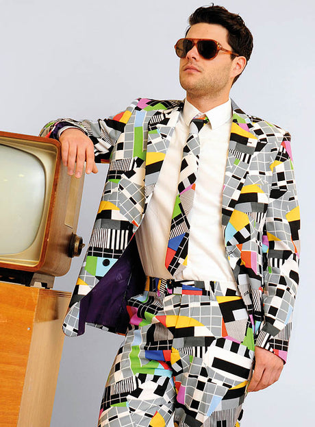 Ein Mann in einem farbenfrohen „Deluxe Testbild“-Anzug von OppoSuits und mit Sonnenbrille lehnt an einem Retro-Fernseher – die ideale Wahl für eine 70er-Jahre-Mottoparty oder als auffälliges Karnevalskostüm.
