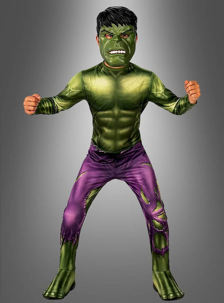 Ein Kind im „Hulk Deluxe“-Kinderkostüm – mit grüner Maske, muskulöser Brust und zerrissener lila Hose – steht mit geballten Fäusten vor einem grauen Hintergrund.