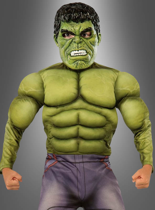 Eine Person trägt das „Hulk Deluxe“-Kinderkostüm, das sich durch einen grünen, muskulösen Look mit geballten Fäusten und einem grimmigen Gesichtsausdruck auszeichnet, und steht vor einem grauen Hintergrund mit Farbverlauf.