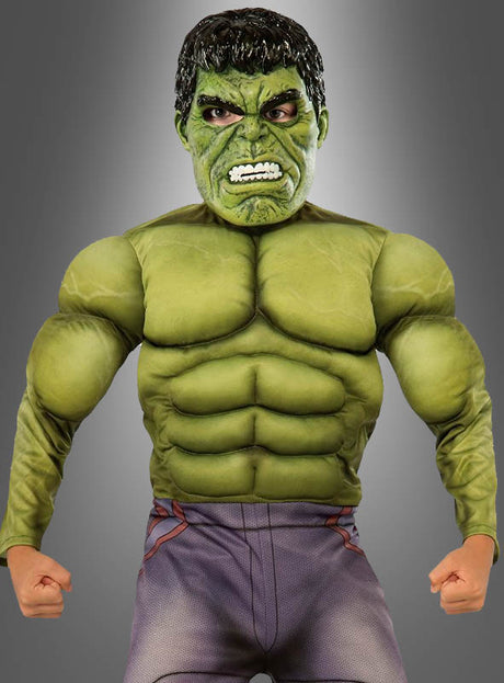 Eine Person trägt das „Hulk Deluxe“-Kinderkostüm, das sich durch einen grünen, muskulösen Look mit geballten Fäusten und einem grimmigen Gesichtsausdruck auszeichnet, und steht vor einem grauen Hintergrund mit Farbverlauf.
