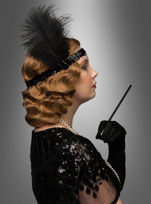 Eine Frau im Profil trägt ein Flapper-Kleid im Stil der 1920er Jahre mit einem gefiederten Stirnband, einer Perlenkette und einem Paillettenkleid und hält vor einem grauen Farbverlaufshintergrund eine Zigarettenspitze in der Hand. Abgerundet wird ihr Look durch die „Premium Luxus Perücke 20er Wasserwelle Frisur Lilly“, die perfekt gestylt ist, um den eleganten Glamour dieser Ära zu evozieren.