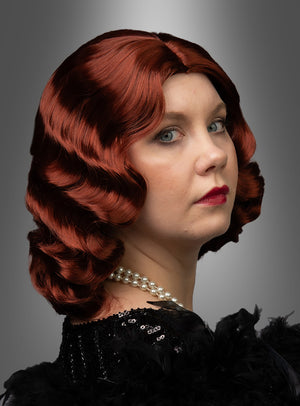 Eine Frau mit welligem, kinnlangem rotem Haar, das im Stil der eleganten „Premium Luxus Perücke 20er Wasserwelle Frisur Lilly“ frisiert ist und an die 1920er Jahre erinnert, trägt ein schwarzes Outfit mit Federbesatz und eine Perlenkette und blickt vor einem grauen Hintergrund mit Farbverlauf leicht über die Schulter.