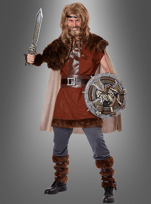 Eine Person im Wikinger-Kostüm „Nordmann“, die ein Schwert und einen verzierten runden Schild in der Hand hält, trägt ein mit Pelz besetztes Outfit sowie braune Stiefel und steht vor einem grauen Hintergrund.