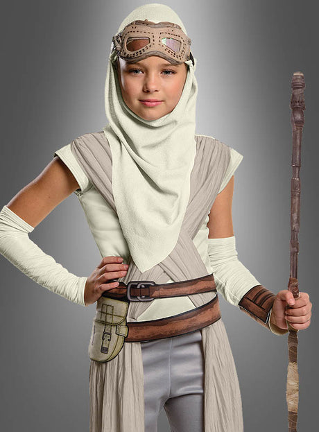 Ein Kind trägt die „Star Wars 7 Rey“-Brille mit Kapuze für Kinder, bestehend aus einem beige-grauen Kostüm mit Kapuze, einer auf der Stirn ruhenden Schutzbrille und einem Stab, und steht vor einem einfarbigen grauen Hintergrund.