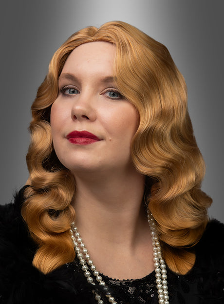 Eine Frau mit welligem blondem Haar, gestylt im Stil der 20er Jahre mit der Premium-Wasserwelle-Perücke „Charlotte“, rotem Lippenstift, schwarzem Outfit, einer Perlenkette und einem Accessoire mit Federn posiert vor einem grauen Hintergrund mit Farbverlauf.