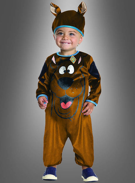 Ein kleines Kind steht lächelnd vor einem grauen Hintergrund und trägt dabei das Scooby-Doo-Hund-Kinderkostüm, ein originales, braunes, lizenziertes Kostüm für Kinder mit Hundeohren und einem aufgedruckten Scooby-Doo-Gesicht.