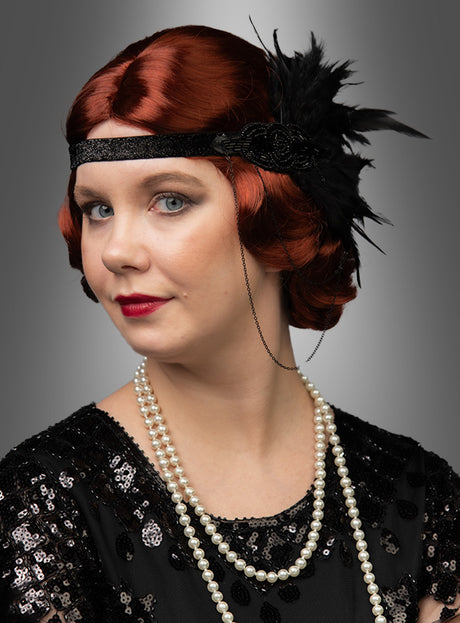 Eine Frau mit kurzen roten Haaren, die im Stil der „Deluxe 20er Wasserwelle Kurzhaar-Frisur Irene Premium“ mit eleganten Wellen im Stil der 1920er Jahre frisiert sind, trägt ein schwarzes Haarreif mit Federn, ein schwarzes Paillettenkleid und eine lange Perlenkette vor einem grauen Hintergrund.