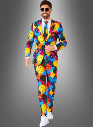 Ein Mann, der den „Bunter Anzug im Smiley-Design“ trägt – einen farbenfrohen Anzug, der mit bunten Smileys verziert ist –, kombiniert mit einem weißen Hemd, einer Krawatte, weißen Sneakers und einer Sonnenbrille, steht vor einem grauen Hintergrund mit Farbverlauf.
