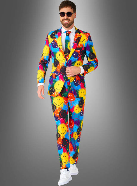 Ein Mann, der den „Bunter Anzug im Smiley-Design“ trägt – einen farbenfrohen Anzug, der mit bunten Smileys verziert ist –, kombiniert mit einem weißen Hemd, einer Krawatte, weißen Sneakers und einer Sonnenbrille, steht vor einem grauen Hintergrund mit Farbverlauf.