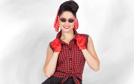 Eine Frau in einem rot-schwarz karierten, ärmellosen Shirt, roten Handschuhen und einem roten Kopftuch mit Polka-Dots, die eine Cat-Eye-Sonnenbrille trägt und vor einem weißen Hintergrund lächelt.