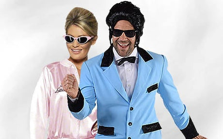 Eine Frau in einer rosa Satinjacke und ein Mann in einem blauen Smoking mit schwarzer Perücke und Sonnenbrille posieren gemeinsam vor einem schlichten Hintergrund.