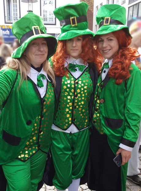 Drei Personen, gekleidet in fröhliche grüne Leprechaun-Kostüme im Stil irischer Kobolde, komplett mit passenden Hüten, mit goldenen Kleeblättern verzierten Westen und Fliegen, stehen gemeinsam da und lächeln für ein Foto bei einer St. Patrick’s Day-Veranstaltung im Freien.
