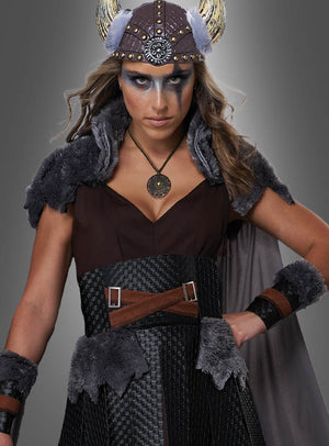 Eine Frau im „Sexy Wikingerin Schildmaid“-Kostüm, das sich durch wikingerinspirierte Details wie Kunstfellakzente, einen gehörnten Helm, auffälliges Make-up und einen runden Anhänger auszeichnet, steht vor einem grauen Hintergrund – ideal für jede Kostümveranstaltung, sei es als Schildmaid oder als Walküre-Karnevalskostüm.