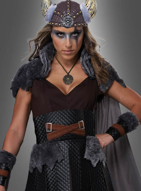 Eine Frau im „Sexy Wikingerin Schildmaid“-Kostüm, das sich durch wikingerinspirierte Details wie Kunstfellakzente, einen gehörnten Helm, auffälliges Make-up und einen runden Anhänger auszeichnet, steht vor einem grauen Hintergrund – ideal für jede Kostümveranstaltung, sei es als Schildmaid oder als Walküre-Karnevalskostüm.