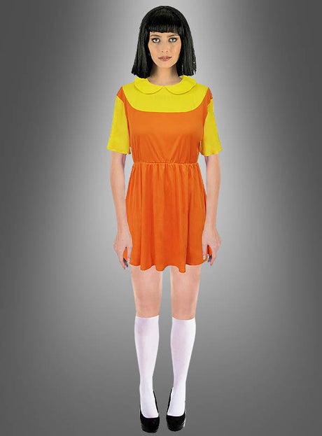 Eine Person trägt das „Kinderspiel Puppe“-Kostüm – „Squad Game“, ein kurzes orange-gelbes Kleid, das vom legendären „Rotes Licht, Grünes Licht“-Kleid inspiriert ist, dazu weiße Kniestrümpfe und schwarze Schuhe; sie steht vor einem grauen Hintergrund mit Farbverlauf.