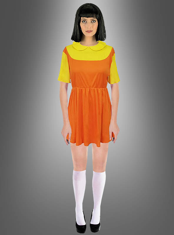 Eine Person trägt das „Kinderspiel Puppe“-Kostüm – „Squad Game“, ein kurzes orange-gelbes Kleid, das vom legendären „Rotes Licht, Grünes Licht“-Kleid inspiriert ist, dazu weiße Kniestrümpfe und schwarze Schuhe; sie steht vor einem grauen Hintergrund mit Farbverlauf.