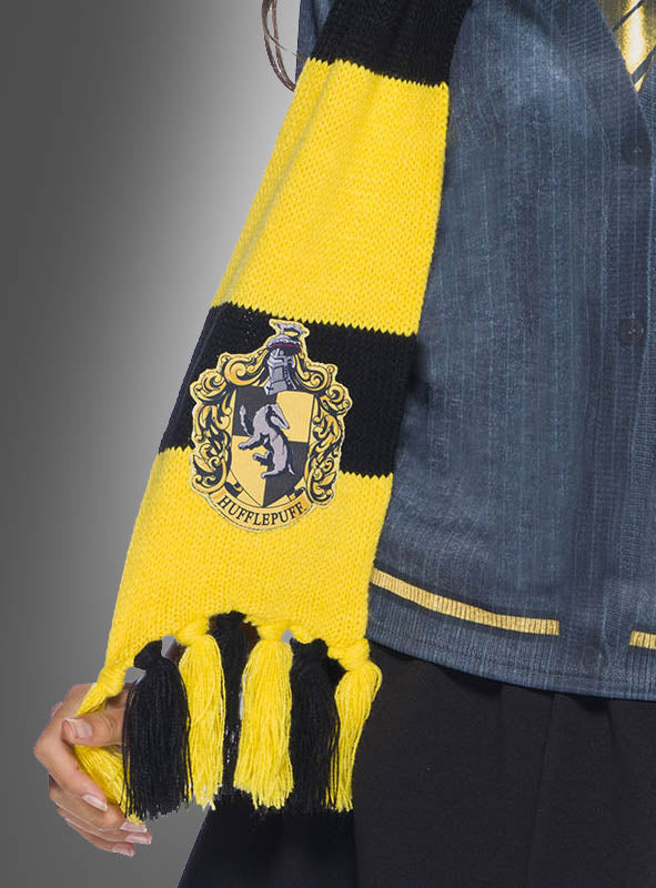 Eine Person, die den gelben Hufflepuff-Schal mit Wappen aus der Harry-Potter-Reihe trägt und diesen mit einer dunklen Jacke und einem Rock als Teil eines offiziellen Harry-Potter-Lizenzkostüms kombiniert hat, steht vor einem grauen Hintergrund.
