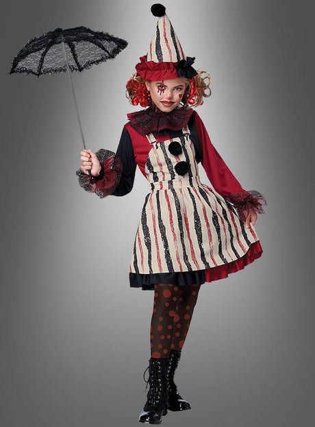 Eine Person, die das Clown-Mädchenkostüm „Columbina“ trägt – bestehend aus einem gestreiften Kleid, einem Rüschenkragen und einem Hut –, hält einen schwarzen Spitzen-Sonnenschirm in der Hand und steht vor einem grauen Hintergrund mit Farbverlauf.