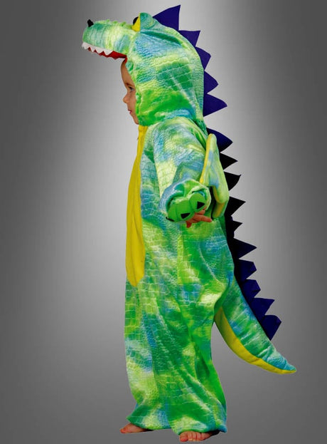 Ein Kind, das das Dinosaurier-Kinderkostüm „Kleiner Drache“ trägt – ein grün-gelbes Kostüm mit Schwanz und Stacheln –, steht barfuß im Profil vor einem grauen Hintergrund.