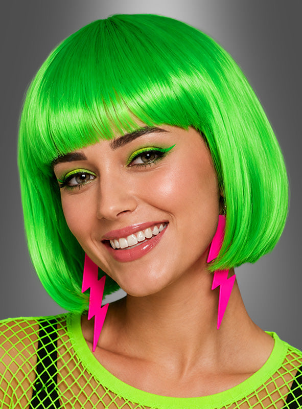 Eine lächelnde Frau mit einer leuchtend grünen, farbenfrohen „Foxy Neon“-Bob-Frisur, neongrünem Augen-Make-up und großen rosa Blitz-Ohrringen, die ein grünes Netz-Top trägt – ein lebhafter 80er-Jahre-Kostüm-Look – vor einem grauen Hintergrund mit Farbverlauf.