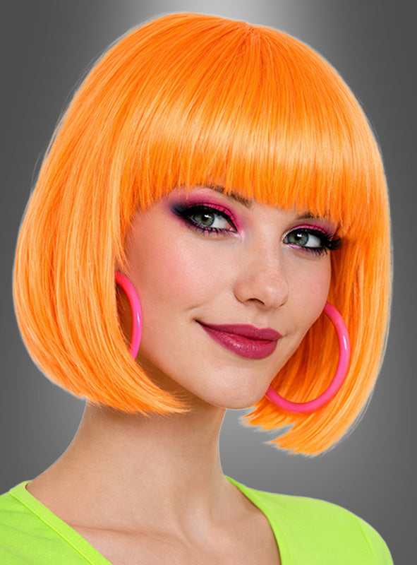 Eine Frau mit einer farbenfrohen „Foxy Neon“-Bob-Frisur in leuchtendem Orange, dazu rosa Creolen, auffälligem rosa und violettem Augen-Make-up sowie einem neongrünen Oberteil posiert selbstbewusst vor einem grauen Hintergrund mit Farbverlauf – ein mühelos stilvoller 80er-Jahre-Kostüm-Look.