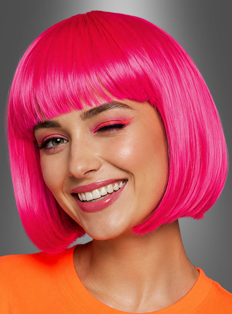Eine Person mit einer leuchtend neonfarbenen „Farbenfrohen Foxy Neon-Bobfrisur“ in Pink und einem orangefarbenen Shirt, die zwinkert, lächelt und rosa Make-up trägt – vor einem grauen Farbverlaufshintergrund – ideal für ein 80er-Jahre-Kostüm.