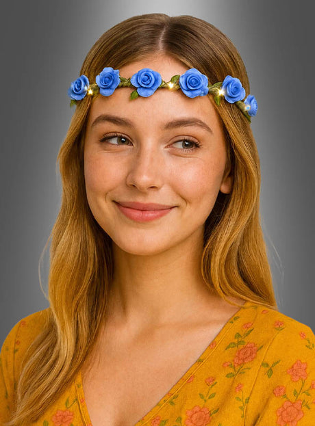 Vor einem grauen Hintergrund mit Farbverlauf lächelt eine Frau und blickt leicht zur Seite. Sie trägt ein gelbes Oberteil mit Blumenmuster und ein Stirnband, das mit blauen Rosen verziert ist. Der zarte Blumen-Haarkranz „Rosen mit LED-Licht“ in ihrem Haar verleiht ihr einen bezaubernden Schimmer und lässt sie so aussehen, als wäre sie für jeden Anlass mit Elfenkostüm-Accessoires bereit.