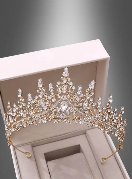 „A Luxus Krone für Königin“, eine goldene Tiara, die mit klaren Edelsteinen verziert ist und an die Eleganz einer Königin-Krone erinnert, wird in einer offenen, hellen Schmuckschatulle vor grauem Hintergrund präsentiert.