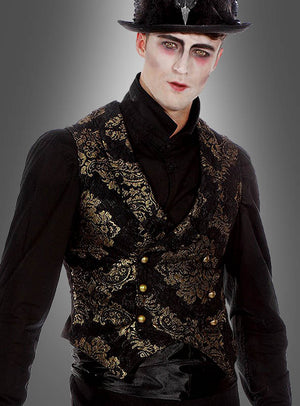 Eine Person, die ein schwarzes Hemd und die „Luxus Gothic Goldbrokat Weste“ – eine aufwendig verzierte schwarz-goldene Weste – trägt, dazu einen schwarzen Hut, blasses Make-up und dunklen Lidschatten, steht vor einem grauen Hintergrund; dieses Ensemble ist von der viktorianischen Mode inspiriert und verbindet klassische Eleganz mit dramatischem Flair.