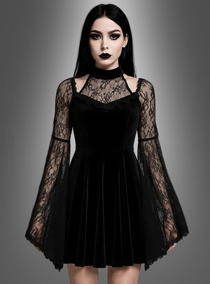 Eine Frau, die das „Schwarzes Gothic Samtkleid Dakaria“ trägt – ein schwarzes Samtkleid mit Spitzen-Glockenärmeln –, steht vor einem grauen Hintergrund mit Farbverlauf und blickt mit ausdruckslosem Gesicht nach vorne.