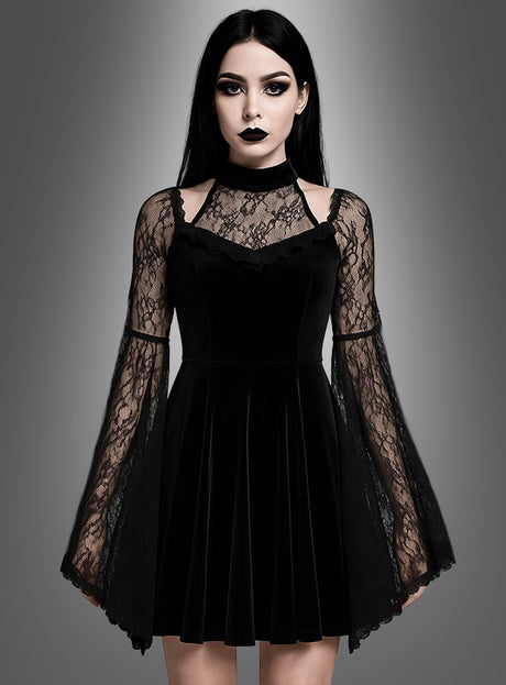 Eine Frau, die das „Schwarzes Gothic Samtkleid Dakaria“ trägt – ein schwarzes Samtkleid mit Spitzen-Glockenärmeln –, steht vor einem grauen Hintergrund mit Farbverlauf und blickt mit ausdruckslosem Gesicht nach vorne.