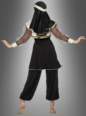 Eine Person steht mit dem Rücken zur Kamera und trägt das Kostüm „Orient Prinzessin Jasmina aus 1001 Nacht“ mit schwarzen und goldenen Details, transparenten Ärmeln, einem Kopfschmuck, einer weiten Hose und hochhackigen Schuhen vor einem grauen Hintergrund mit Farbverlauf.
