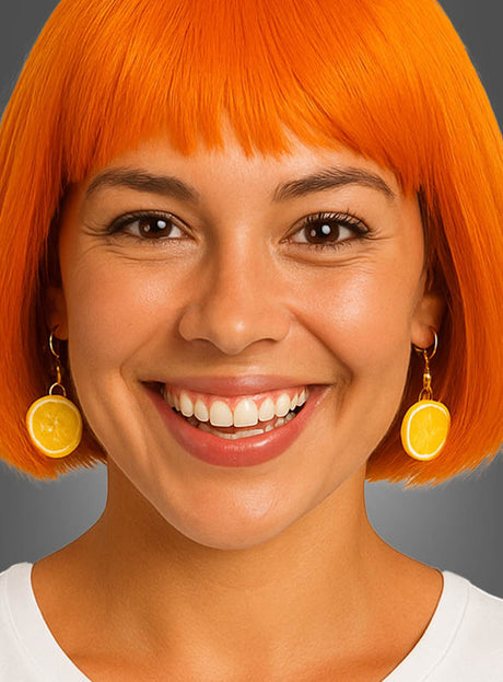 Eine Frau mit einem leuchtend orangefarbenen Bob-Haarschnitt lächelt; sie trägt auffälligen Ohrschmuck – Ohrringe in Form einer Zitronenscheibe oder einer ganzen Zitrone – und ein weißes Hemd, vor einem grauen Hintergrund.