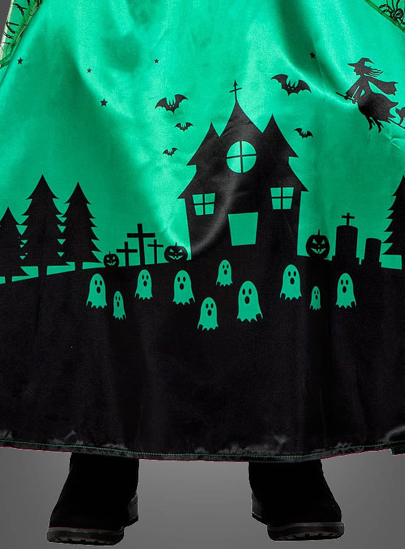 Das lange grüne Hexenkleid für Kinder „Langes Hexenkleid grün für Kinder“ ist mit schwarzen Silhouetten eines Spukhauses, Fledermäusen, einer Hexe, Bäumen, Kürbissen, Grabsteinen und Geistern verziert – perfekt für jedes Halloween-Hexenkostüm. Unter dem Kleid lugen schwarze Stiefel hervor.