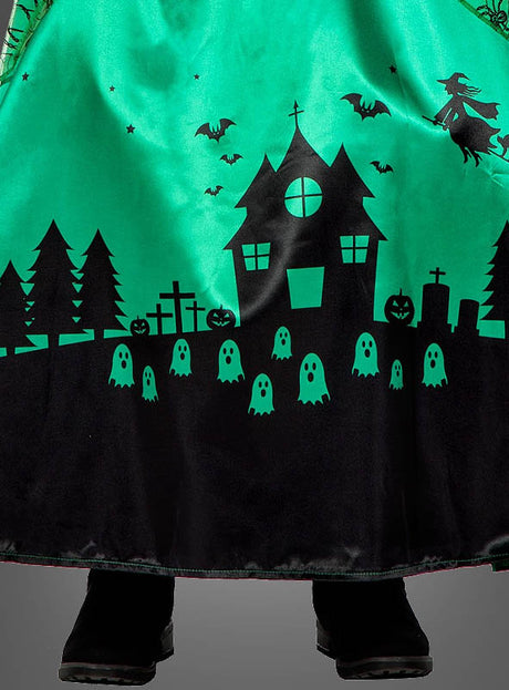 Das lange grüne Hexenkleid für Kinder „Langes Hexenkleid grün für Kinder“ ist mit schwarzen Silhouetten eines Spukhauses, Fledermäusen, einer Hexe, Bäumen, Kürbissen, Grabsteinen und Geistern verziert – perfekt für jedes Halloween-Hexenkostüm. Unter dem Kleid lugen schwarze Stiefel hervor.