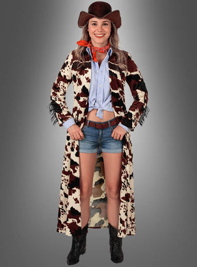 Vor einem grauen Hintergrund steht eine Frau und lächelt. Sie trägt einen Cowboyhut, einen roten Schal, ein blaues Hemd, Jeansshorts, schwarze Stiefel und den „Cowgirl Mantel Kuhfell Design“ – einen langen Staubmantel im klassischen Western-Look mit Kuhmuster.