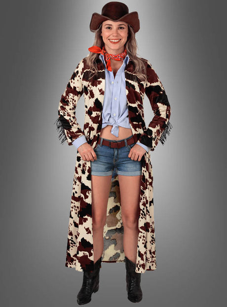 Vor einem grauen Hintergrund steht eine Frau und lächelt. Sie trägt einen Cowboyhut, einen roten Schal, ein blaues Hemd, Jeansshorts, schwarze Stiefel und den „Cowgirl Mantel Kuhfell Design“ – einen langen Staubmantel im klassischen Western-Look mit Kuhmuster.