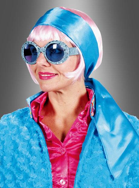 Eine Frau mit einer großen blauen Sonnenbrille, einem blauen Haarband-Halstuch aus Satin als Kopftuch, einer rosa Perücke, einer rosa Bluse und einer blauen Strukturjacke vor grauem Hintergrund – perfekt für eine 70er-Jahre-Party.