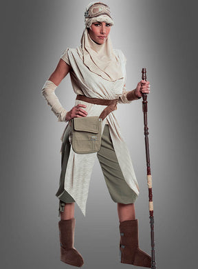 Eine Person, die das „STAR WARS Rey Super Deluxe“-Kostüm trägt – ein beige- und hellbraunes Damen-Filmkostüm im Wüstenstil –, steht mit einem Stab, einer Tasche und einer Kopfbedeckung vor einem grauen Hintergrund und verkörpert damit den ikonischen Look von Rey aus „Star Wars“.